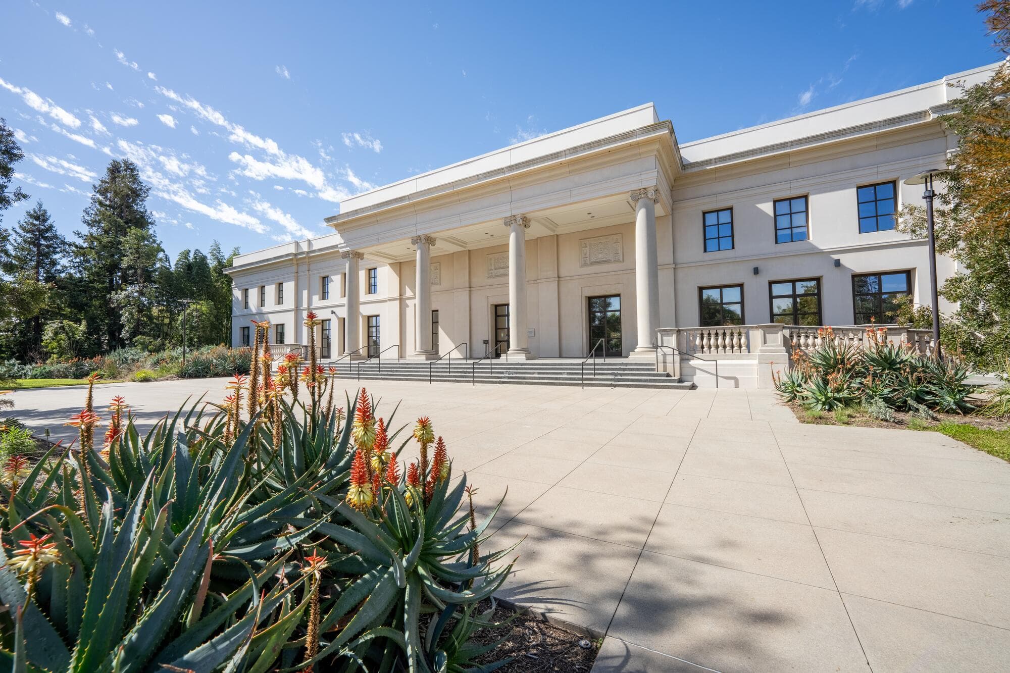A large white building with a concrete courtyard surrounded by gardens.