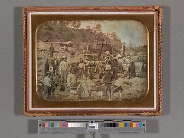 Mining scene of a large group of miners posed while working on a rocky riverbed