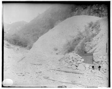 An unidentified place showing a pile of rocks next to a medium sized wood box in front of a small hill amidst the mountains
