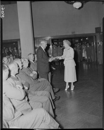 People seated and standing in large room while a man and a woman exchange books