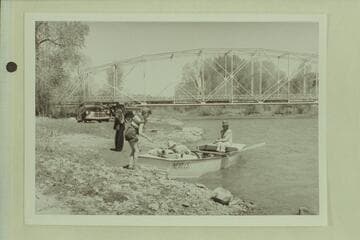Final stowage of the punt before start of the Dolores River traverse.  Bridge below Dolores, Colorado.  Becky Walker stands near the boat.  Marston is stowing dunnage under the deck and Margaret Marston sits on the boat
