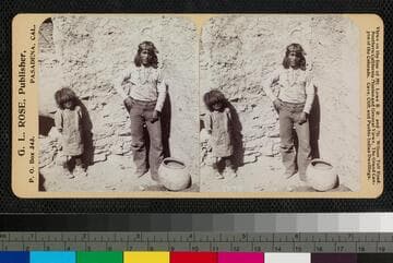 A man and a child stand against an adobe wall in an unidentified pueblo