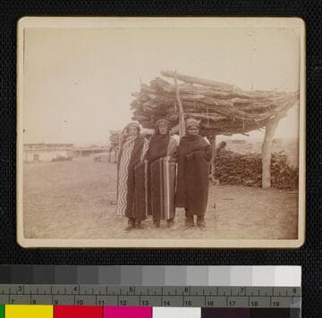 Three Indians, possibly Apache or Yuma, standing in front of wood pile, wearing blankets and head wraps