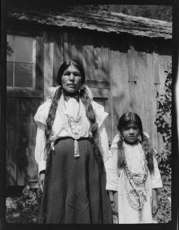 An unidentified Native American woman and girl, wearing necklaces of dentalia, standing in front of house