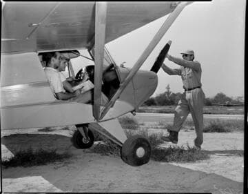 Two people sitting in cockpit of Piper Cub airplane while man turns the prop to start the engine by hand