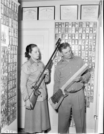 Man and woman in room filled with framed awards and medals for marksmanship. Woman is holding a sharpshooters rifle and man is holding a blank un