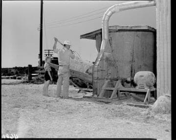 Farmer feeding bales of hay into shredder with daughter standing by