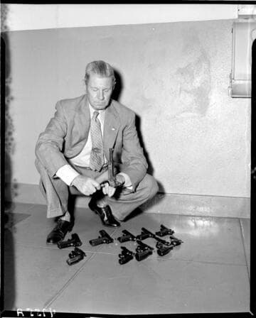 Man kneeling holding German Lueger with a variety of pistols (revolvers, automatics and a derringer) spread out on the floor beside him