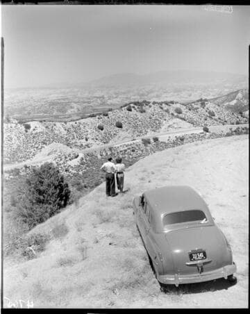 Man and woman standing by their car on a ridge overlooking valley below with mountains in background