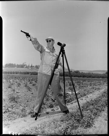 Man aiming pistol in field with spotting scope on tripod next to him