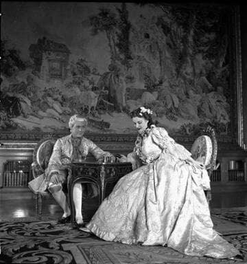 Couple in period costume seated at a small table in the library of the Huntington residence with a Beauvais tapestry in the background